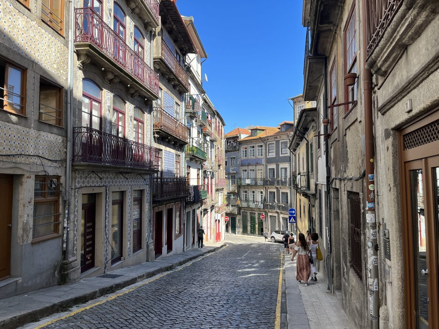 A narrow cobbled street in Porto between tall houses with iron balconies under a blue sky